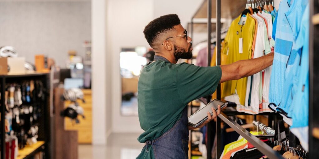 A retail employee using a tablet to count the inventory of clothes on a shelf.